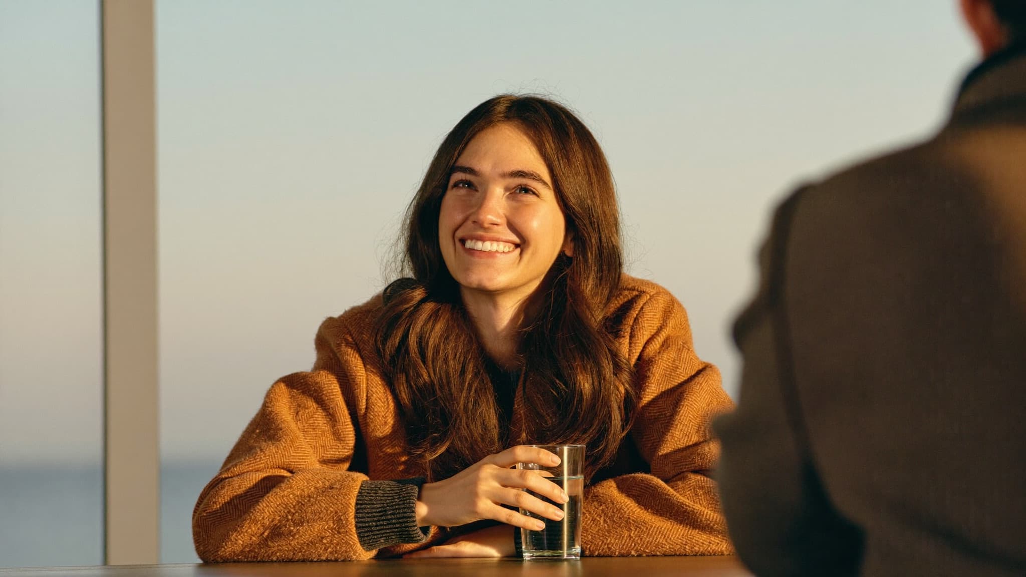 A woman smiling in golden-hour light during a conversation across a table, a glass of water in her hand