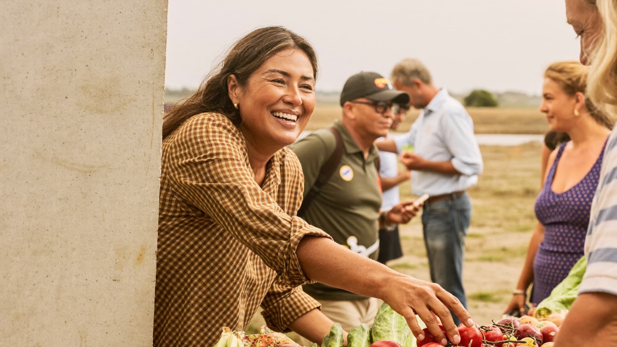 A woman laughing as she reaches over a farm-stand table of tomatoes and greens, other shoppers talking behind her in warm evening haze