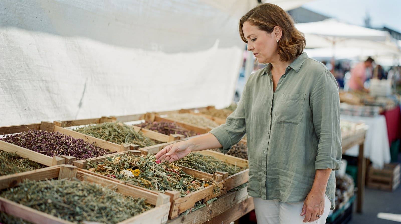 A woman in a green shirt examines dried herbs at an outdoor market stall.