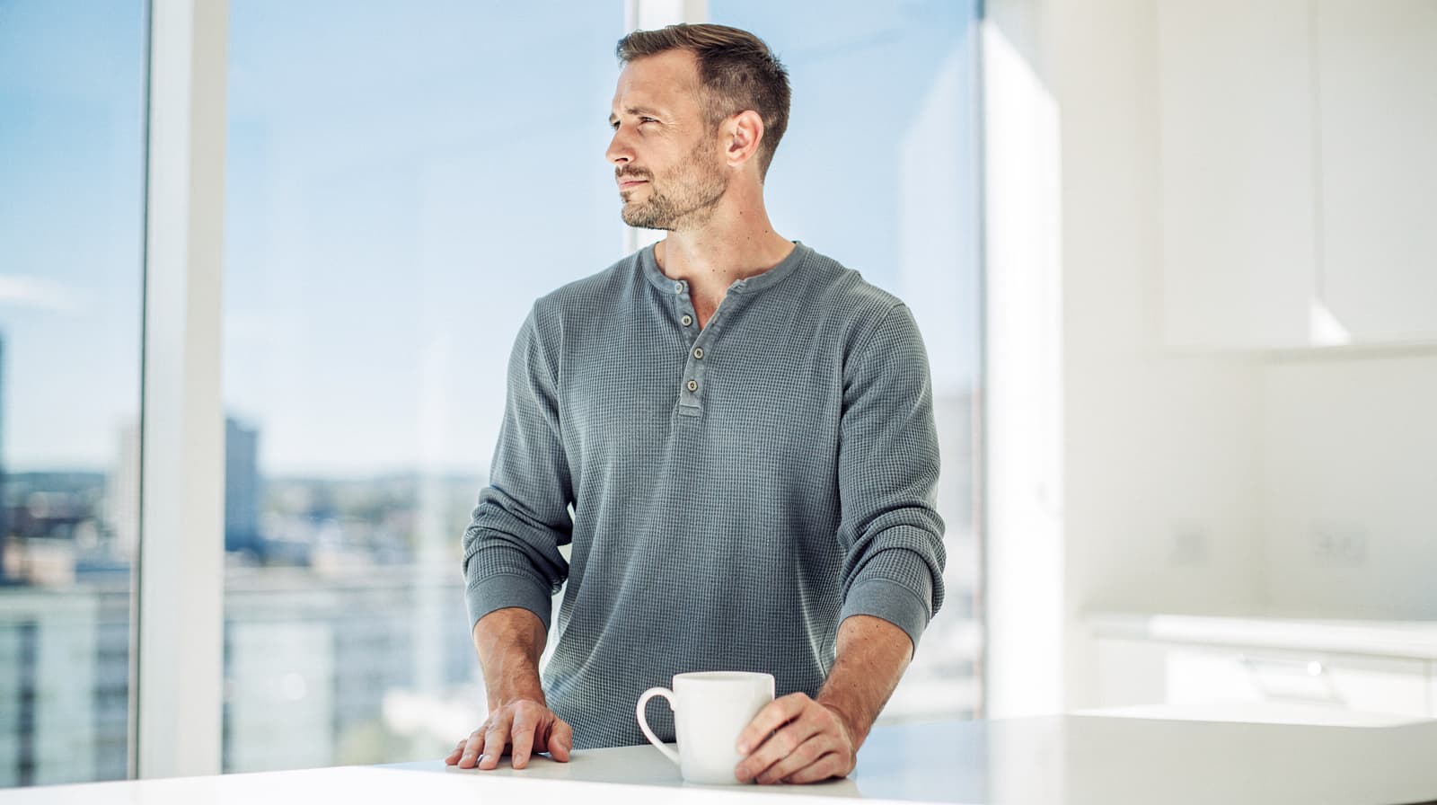 A man in a gray shirt stands at a white counter with a mug, looking out a window in a bright room.