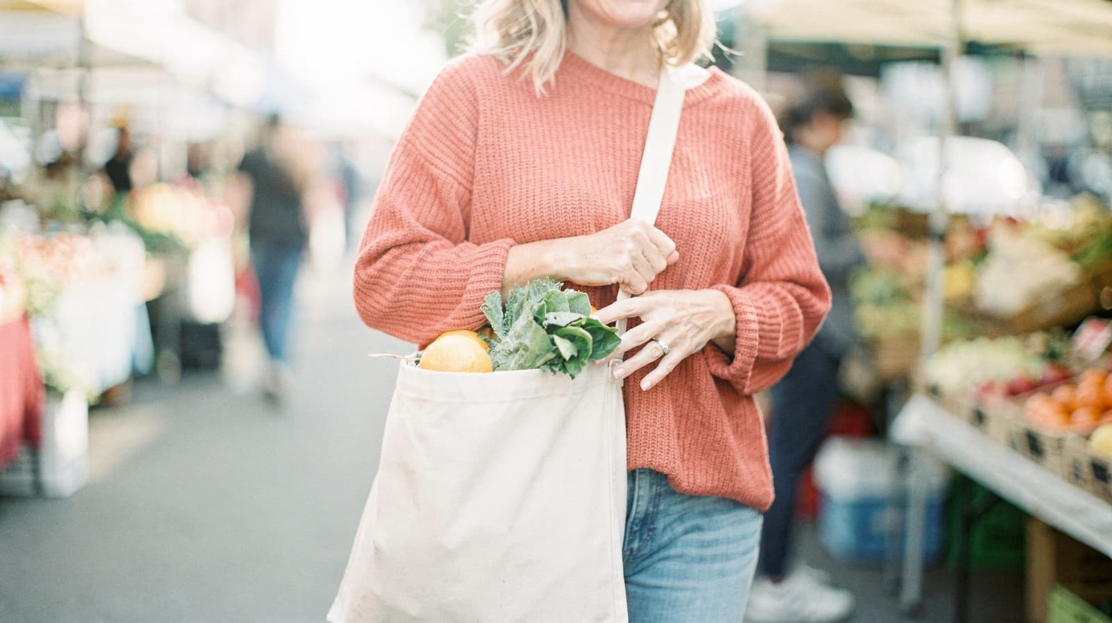 A woman in a red sweater carries a tote bag with produce at an outdoor market.