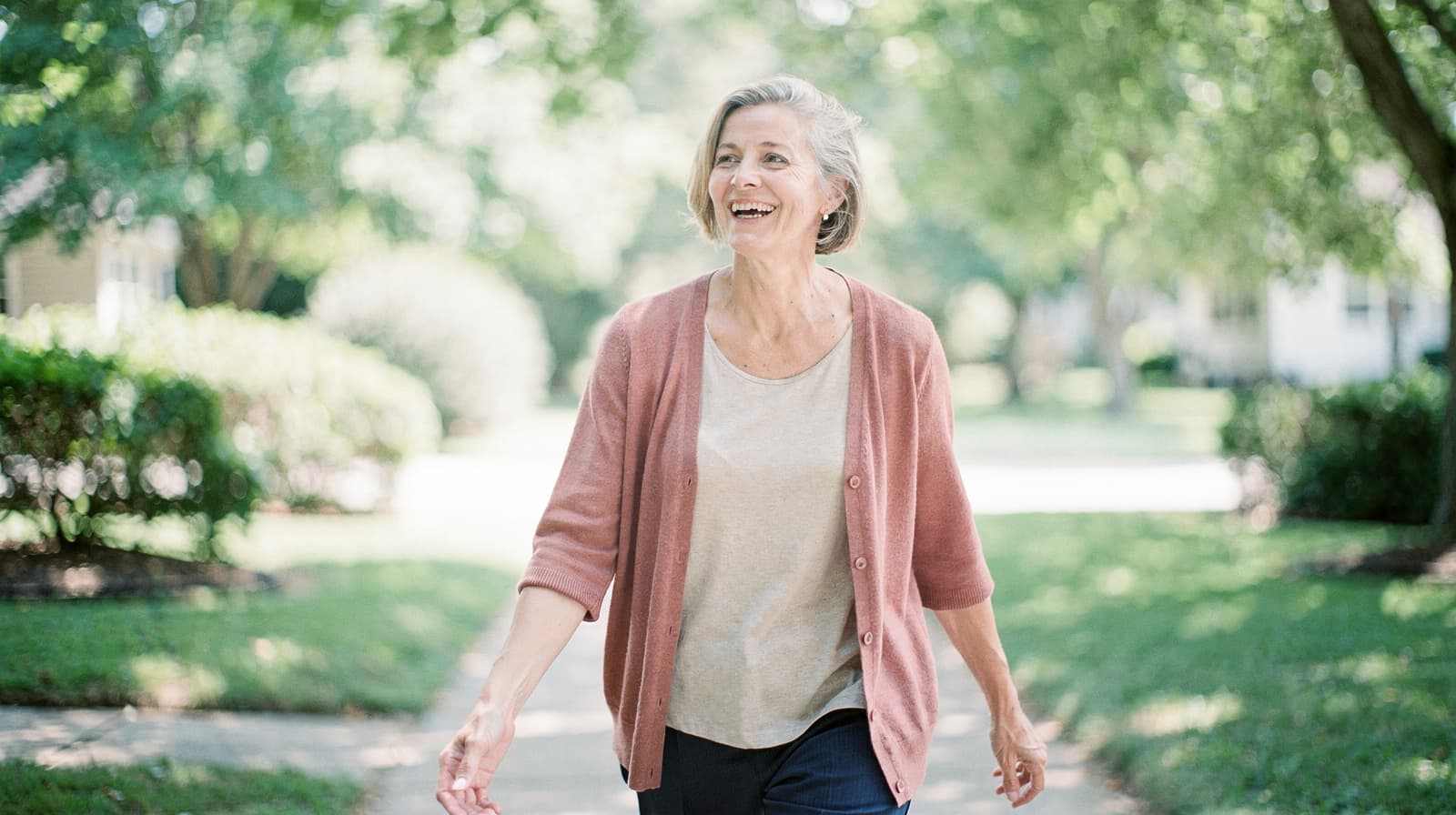 A woman with short gray hair walks along a tree-lined path, smiling and wearing a pink cardigan and beige top.