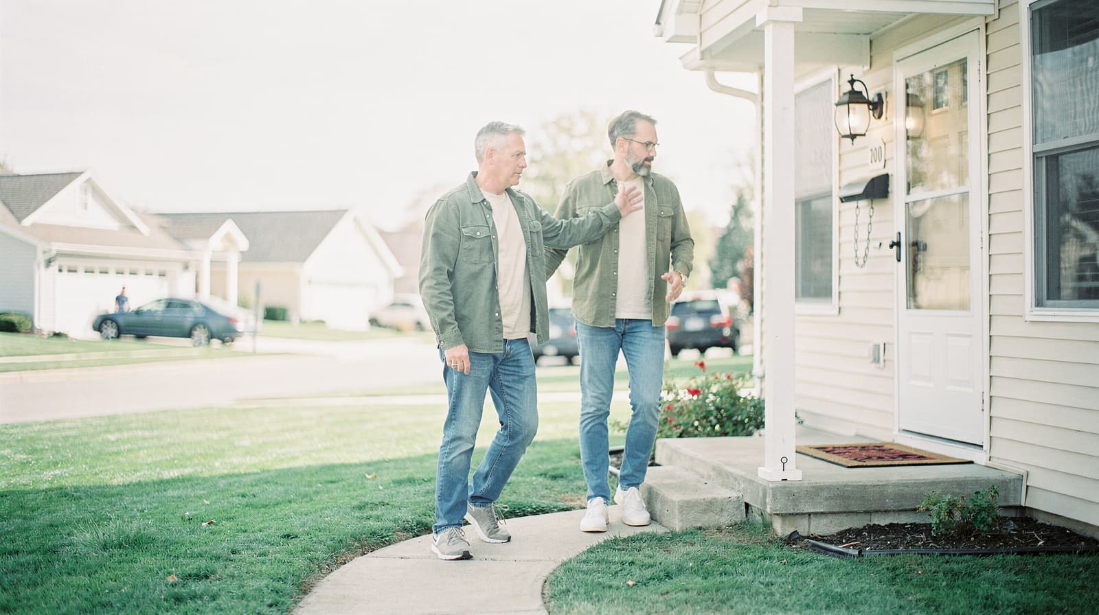 Two men walk on a sidewalk towards a house in a suburban neighborhood.