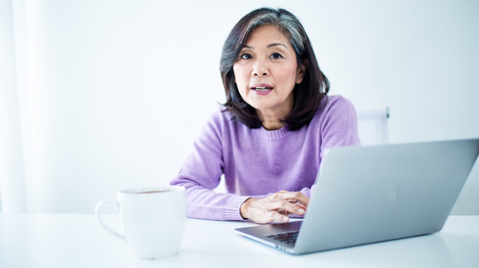 A woman in a purple sweater sits at a table with a laptop and a white mug.