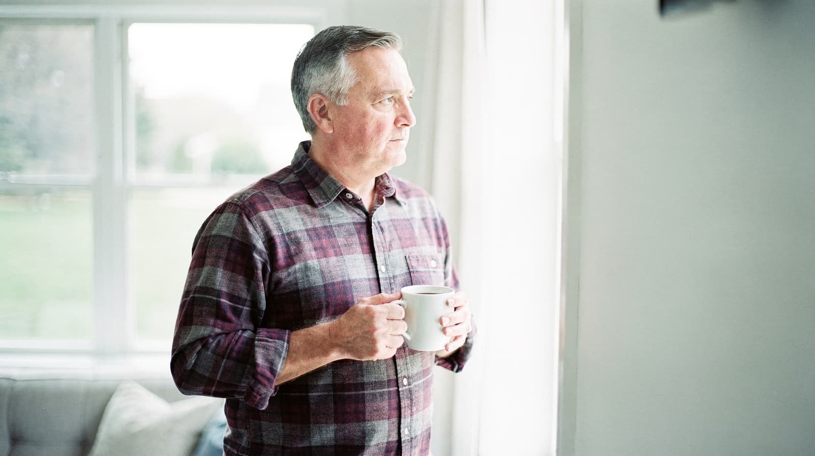 A man in a plaid shirt holds a mug while looking out a bright window in a living room.