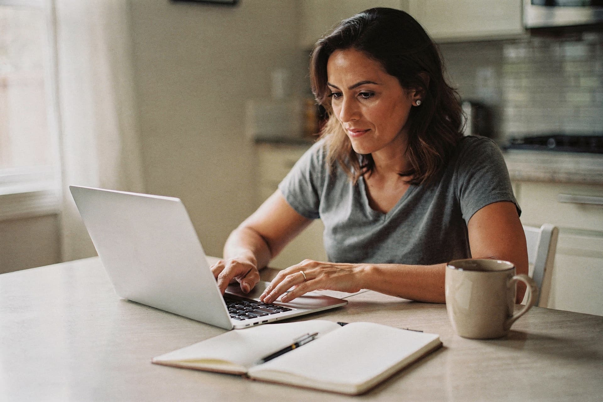 A woman sits at a kitchen table using a laptop with a notebook and mug nearby.