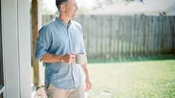 A man in a light blue shirt holds a mug while standing outside near a wooden fence on a sunny day.