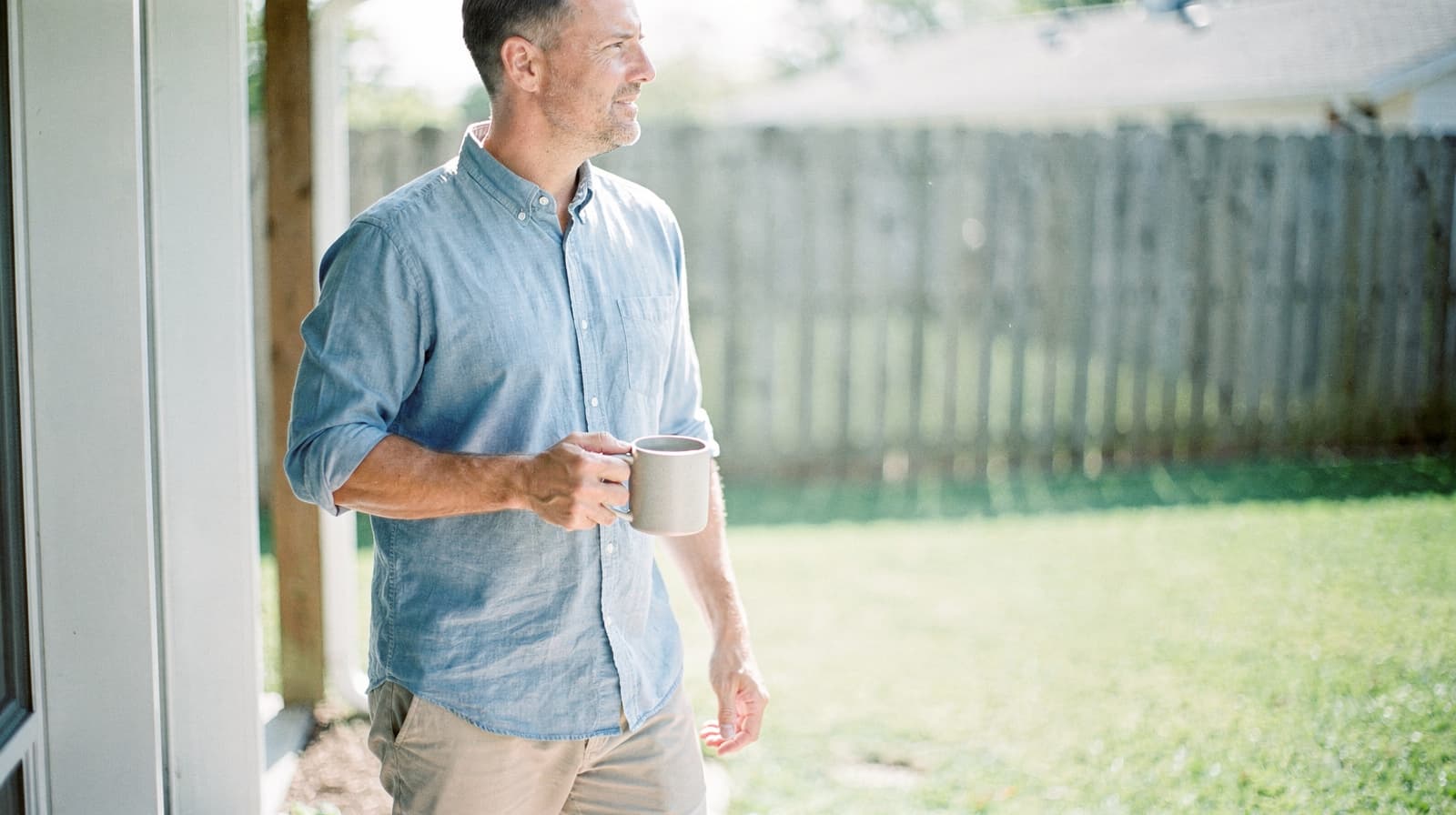A man in a light blue shirt holds a mug while standing outside near a wooden fence on a sunny day.