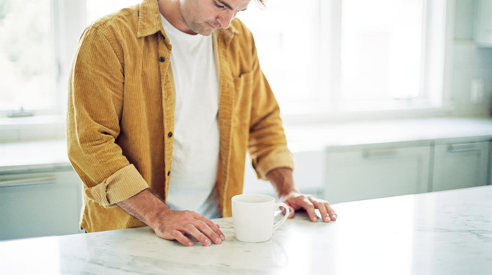 A man in a yellow shirt stands at a kitchen counter with a white mug.