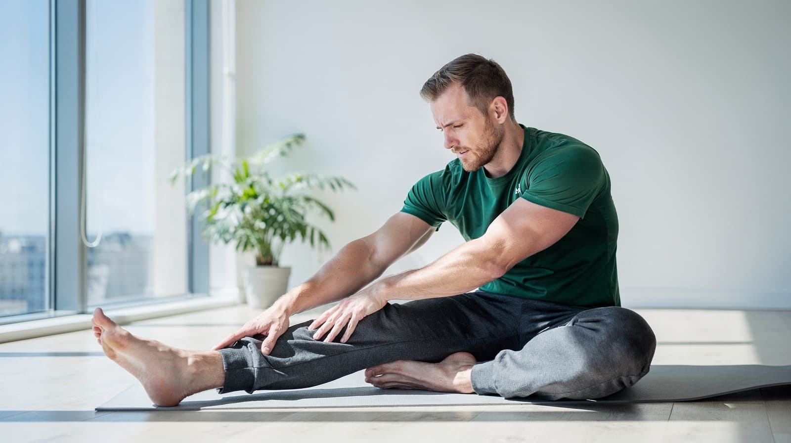 A man in a green shirt stretches on a yoga mat in a bright room with a large window and a potted plant.