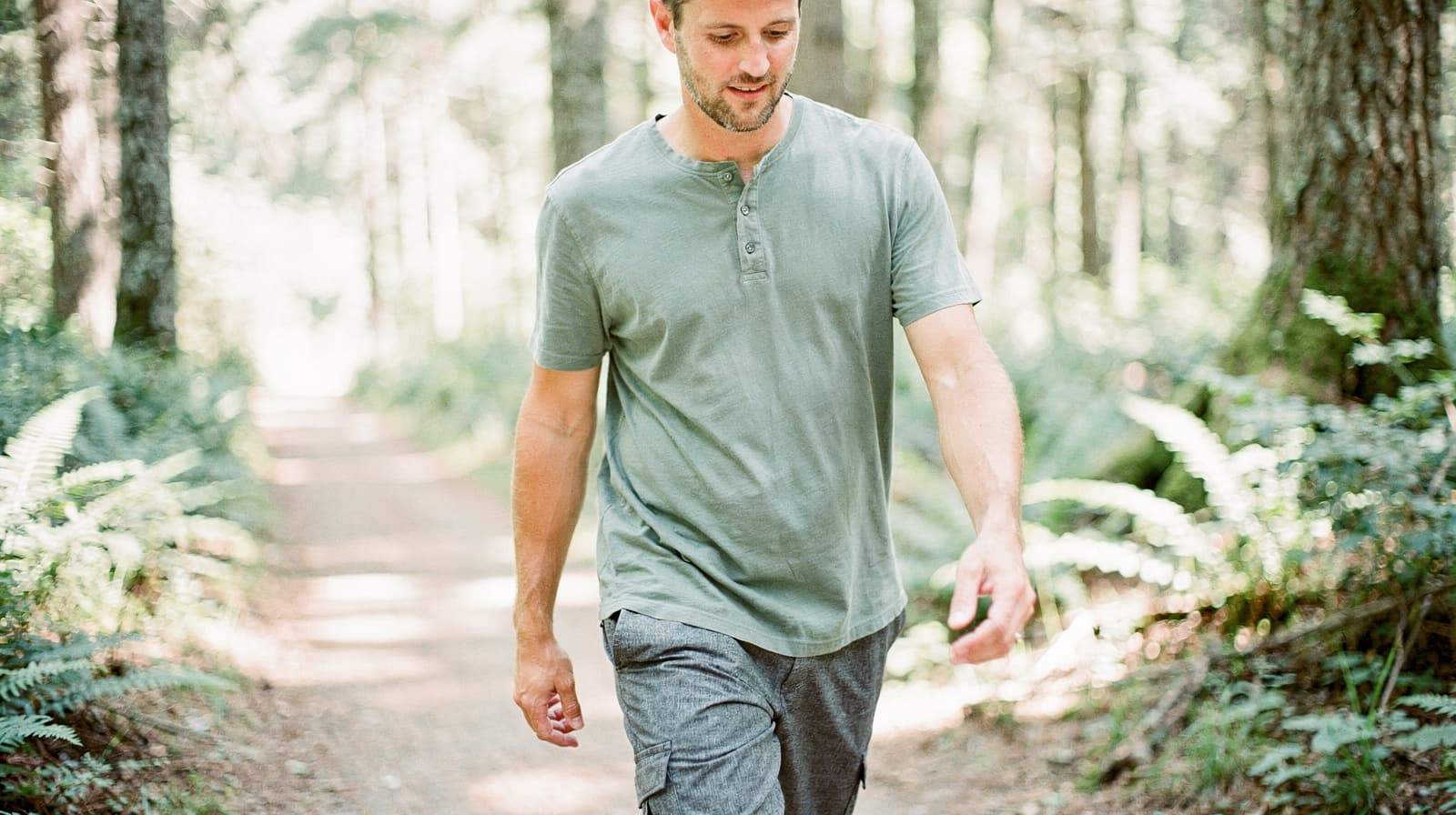 A man walks along a forest path surrounded by trees and ferns.