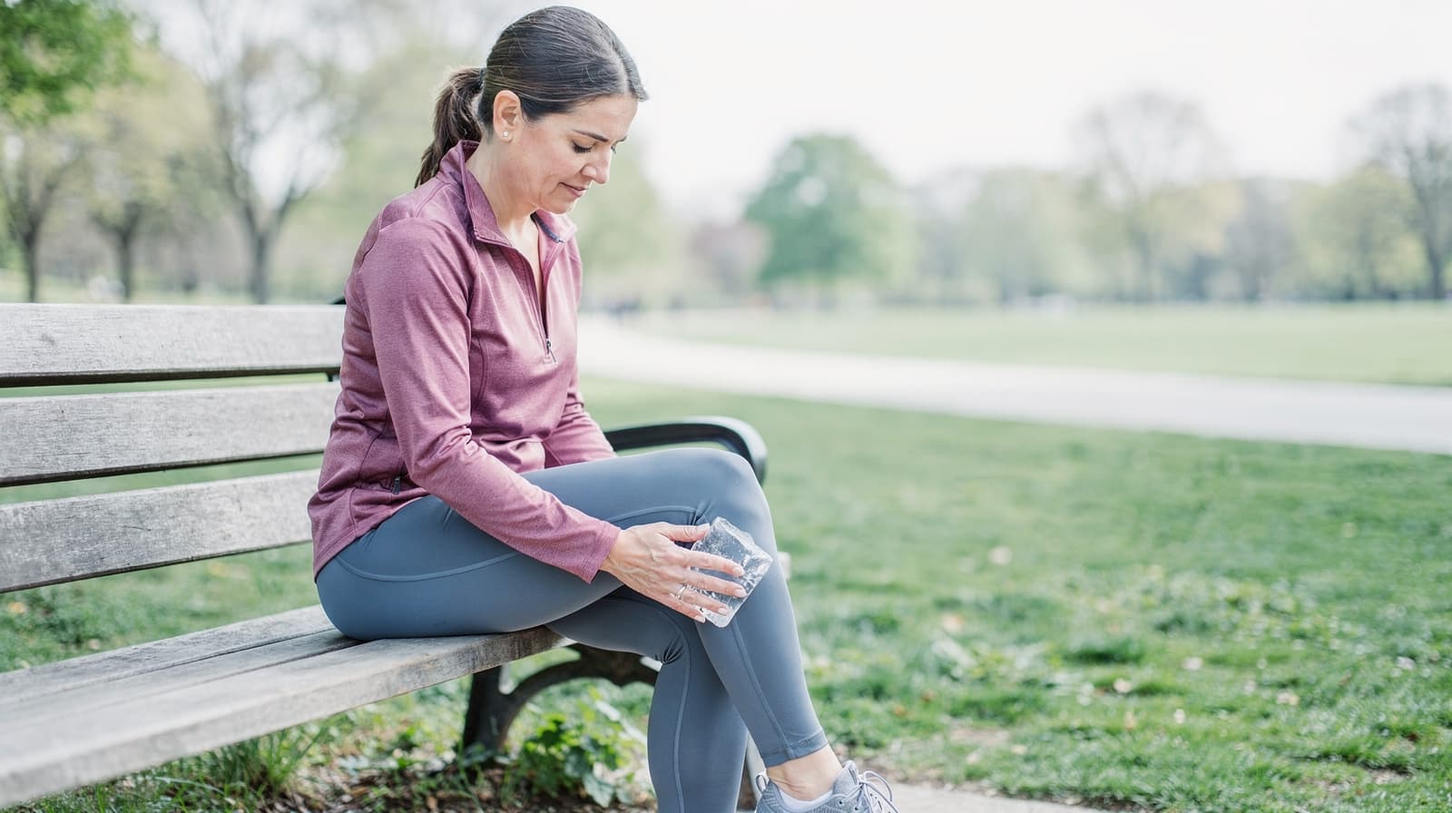 A woman sits on a park bench holding an ice pack on her knee.