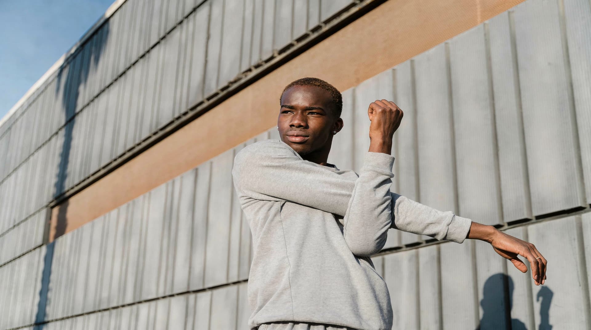 A person in a gray sweatshirt stretches their arm in front of a large, textured wall outdoors.
