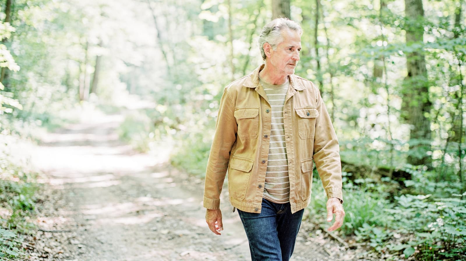 A man in a tan jacket walks along a forest path surrounded by trees.
