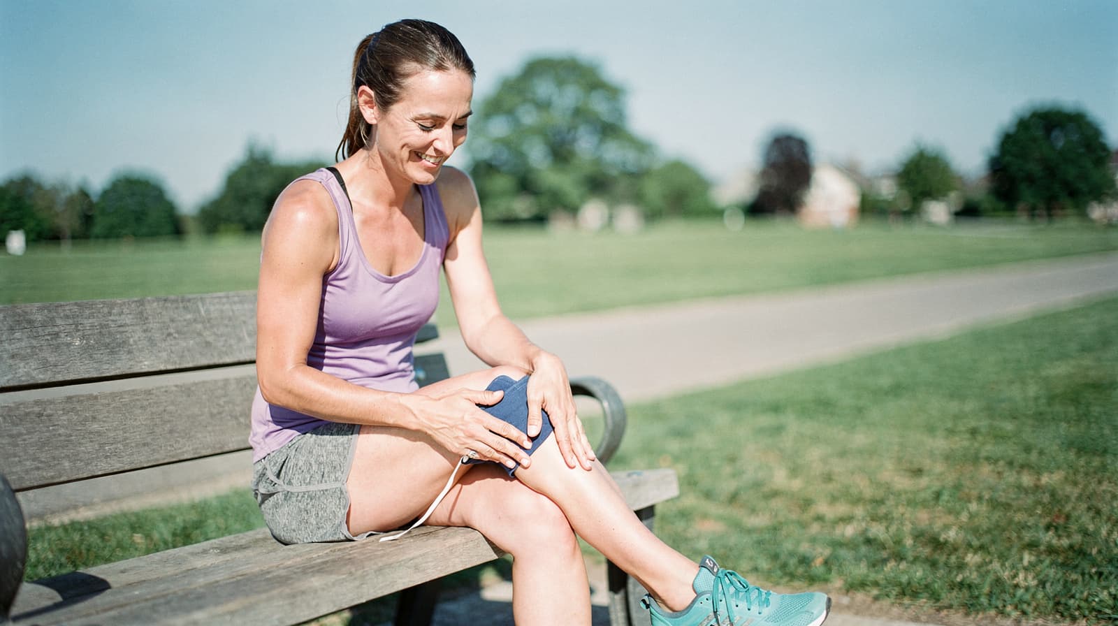 A woman in athletic wear sits on a park bench, smiling and holding an ice pack on her knee.