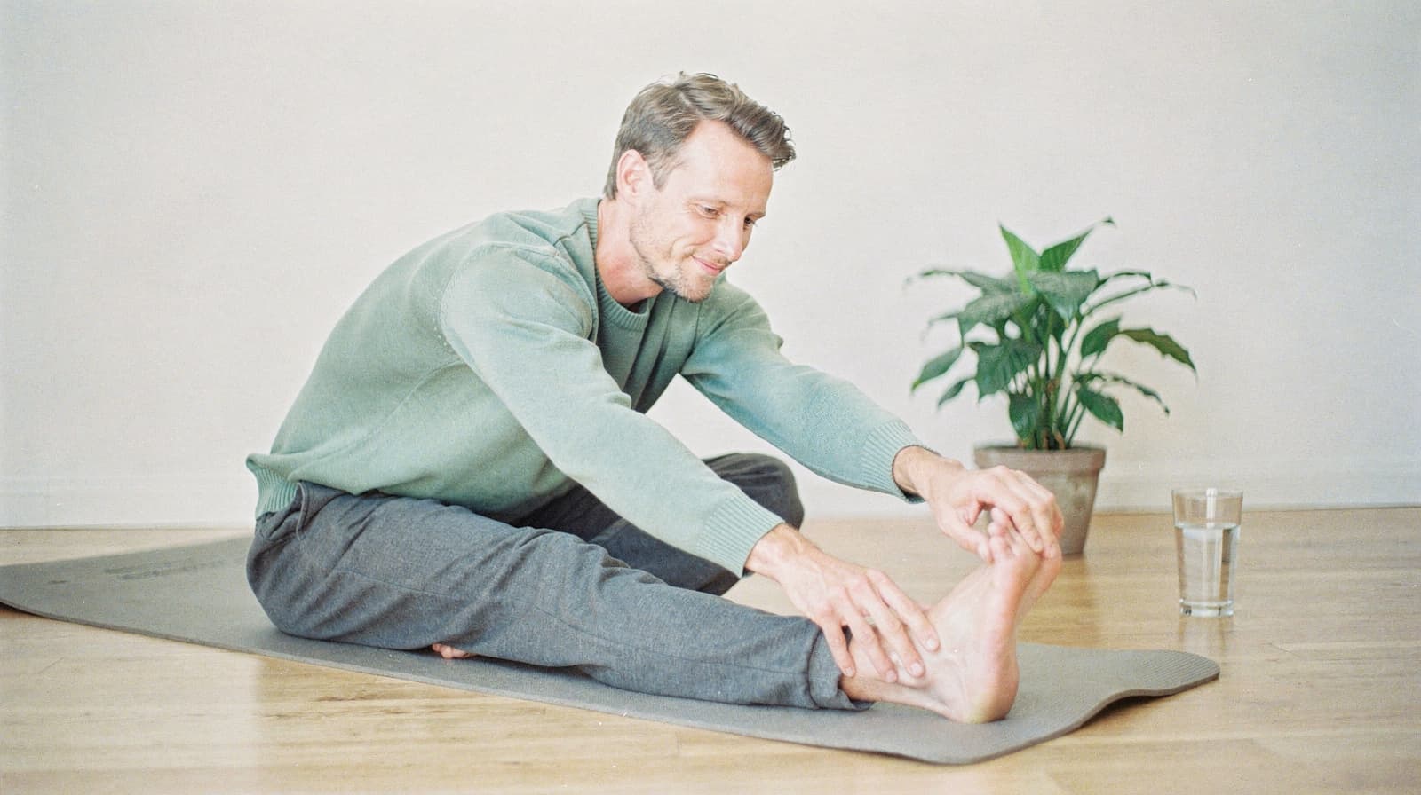 A man sitting on a yoga mat stretches forward to touch his toes, with a potted plant and a glass of water nearby.