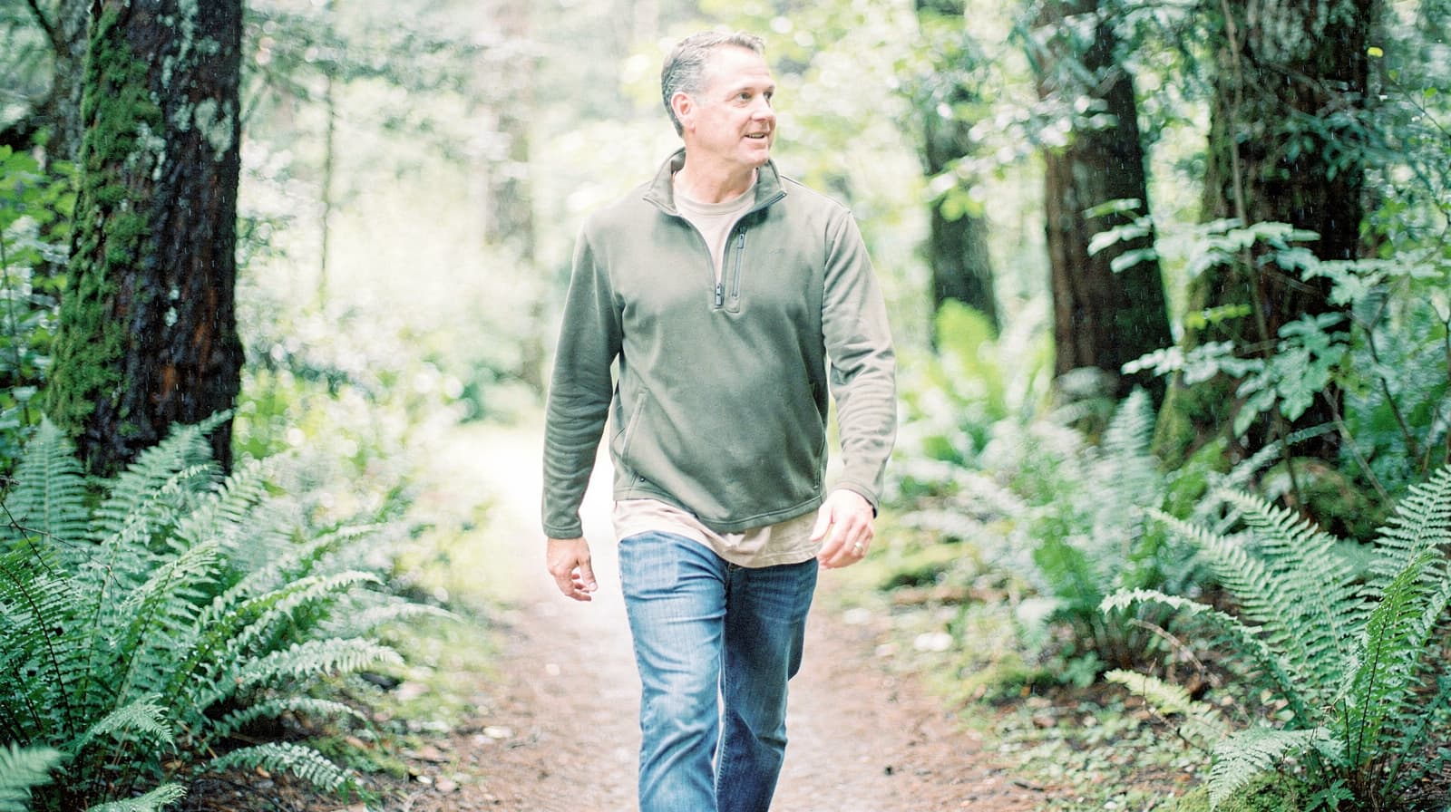A man walks along a forest path surrounded by ferns and trees.