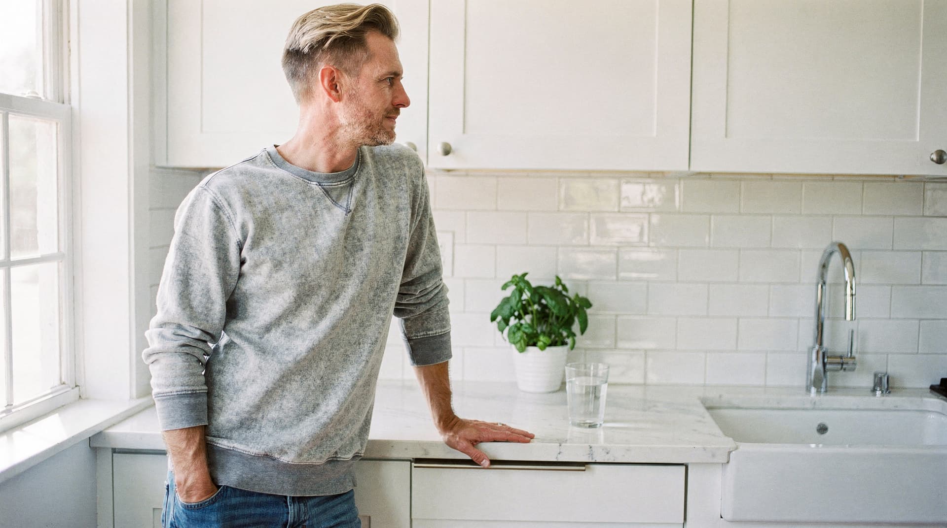 A man in a gray sweater stands in a bright kitchen next to a counter with a potted plant and a glass of water.