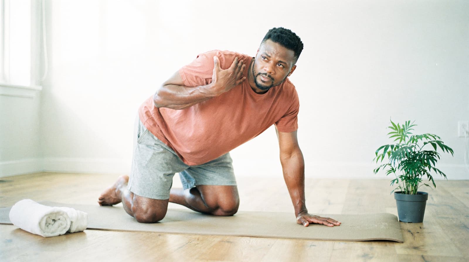 A man in a red shirt and gray shorts kneels on a yoga mat in a bright room with a small plant and a rolled towel nearby.