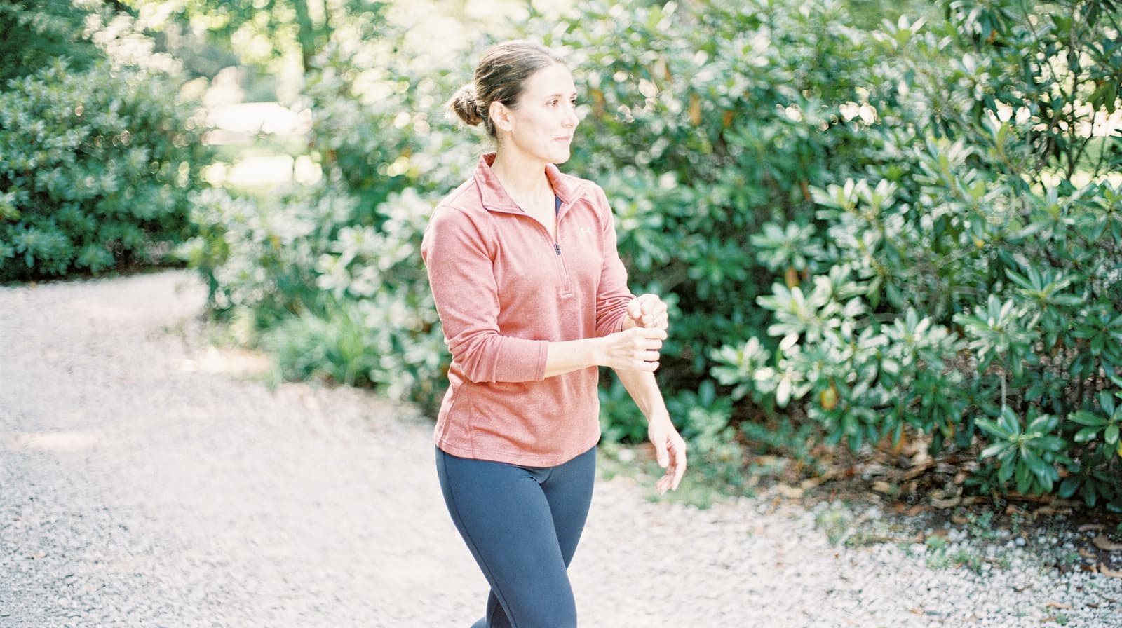 A woman in a red long-sleeve shirt walks on a gravel path surrounded by green foliage.