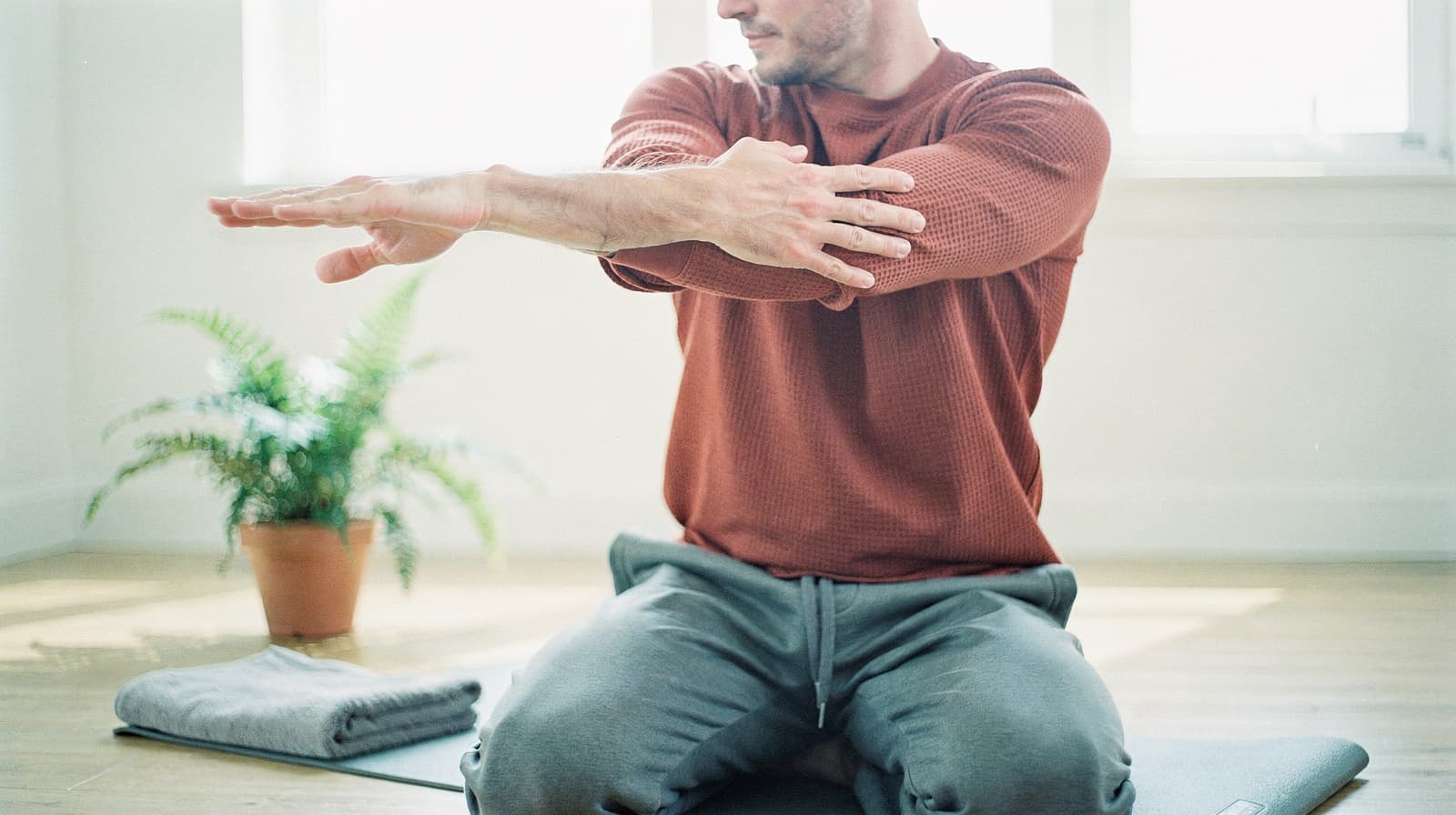 A man in a red sweater stretches his arms while sitting on a yoga mat in a bright room with a potted plant nearby.