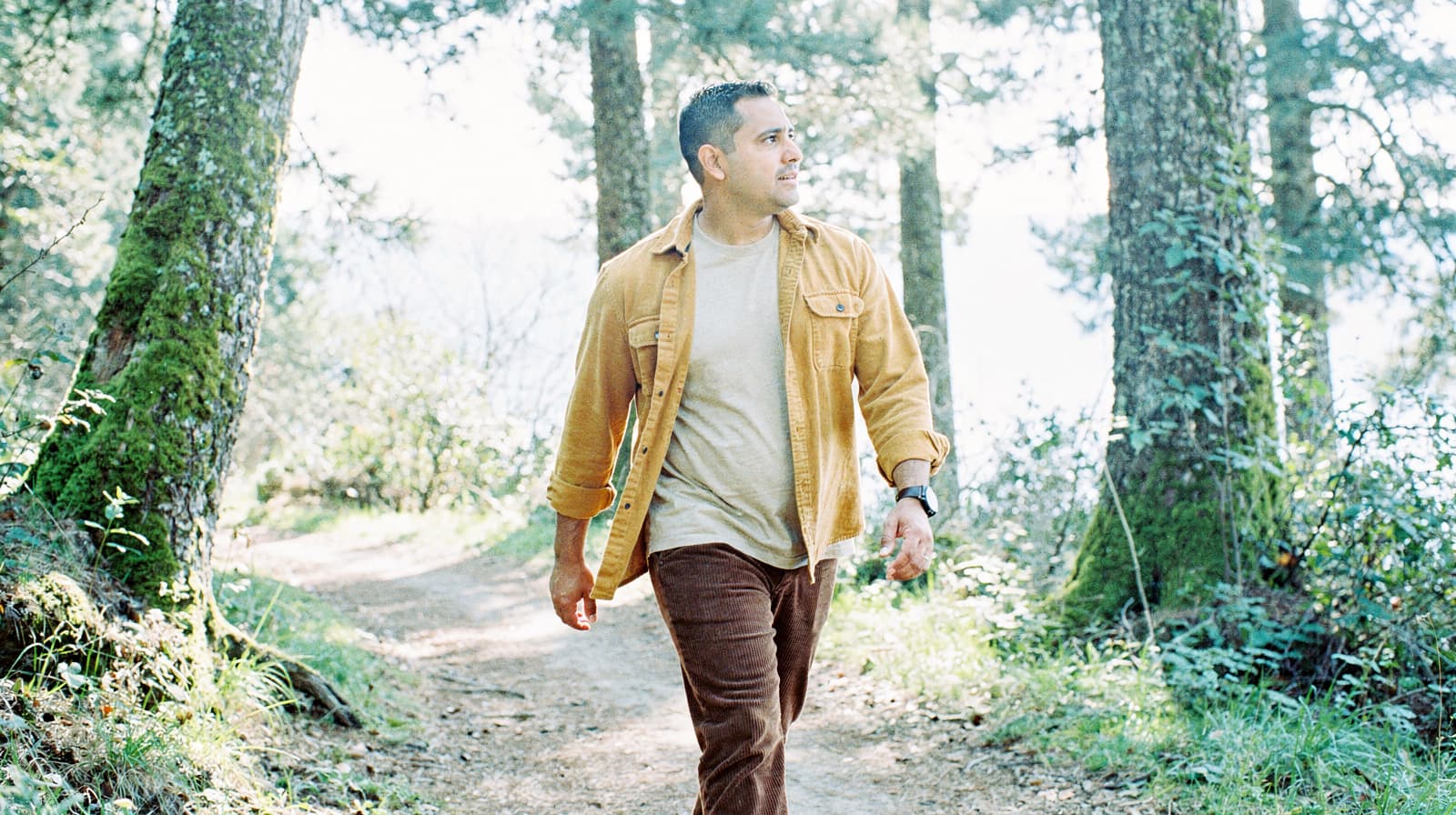 A man walks along a forest path surrounded by tall trees and greenery.