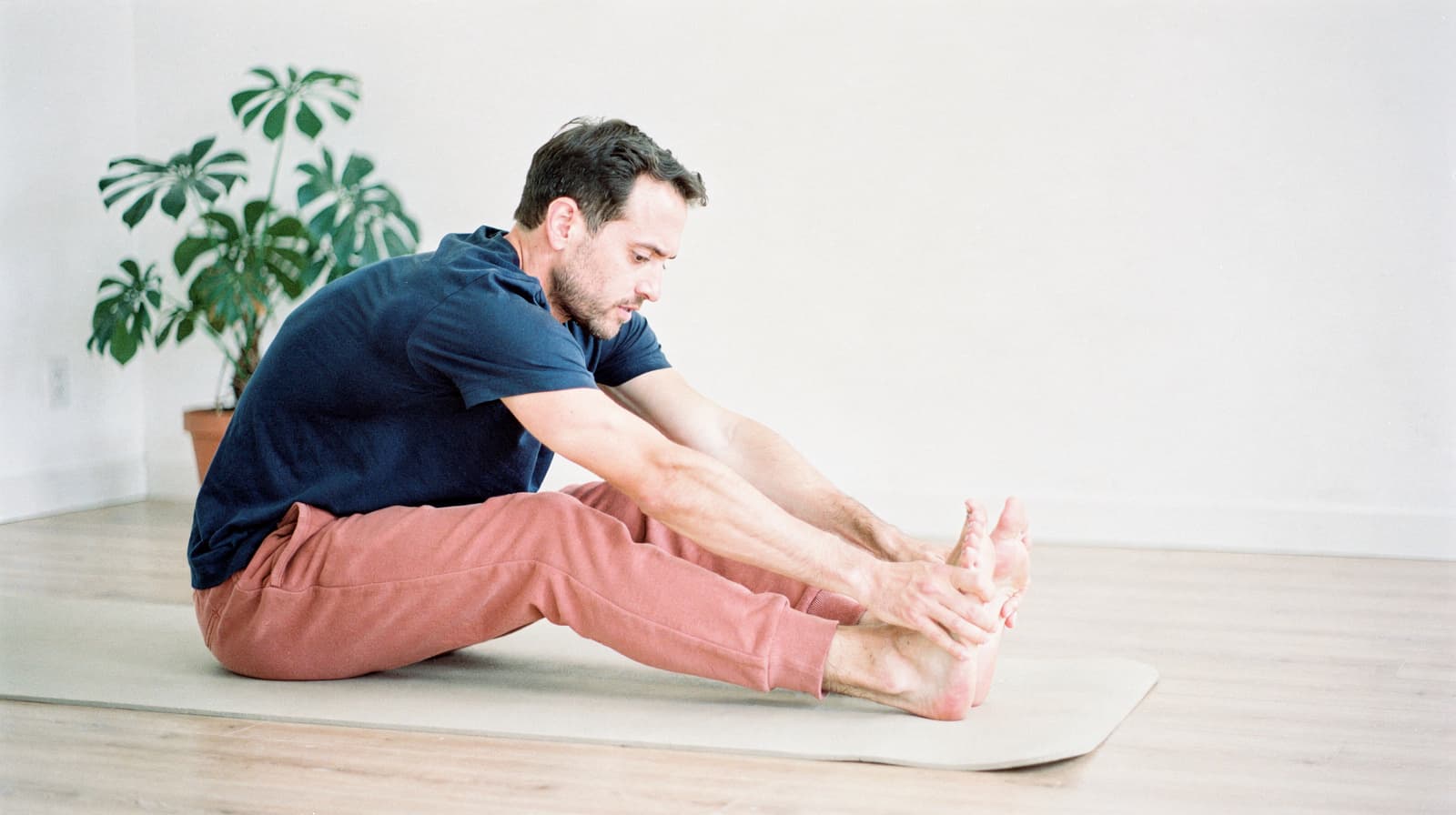 A man sitting on a yoga mat in a bright room stretches forward to touch his toes.