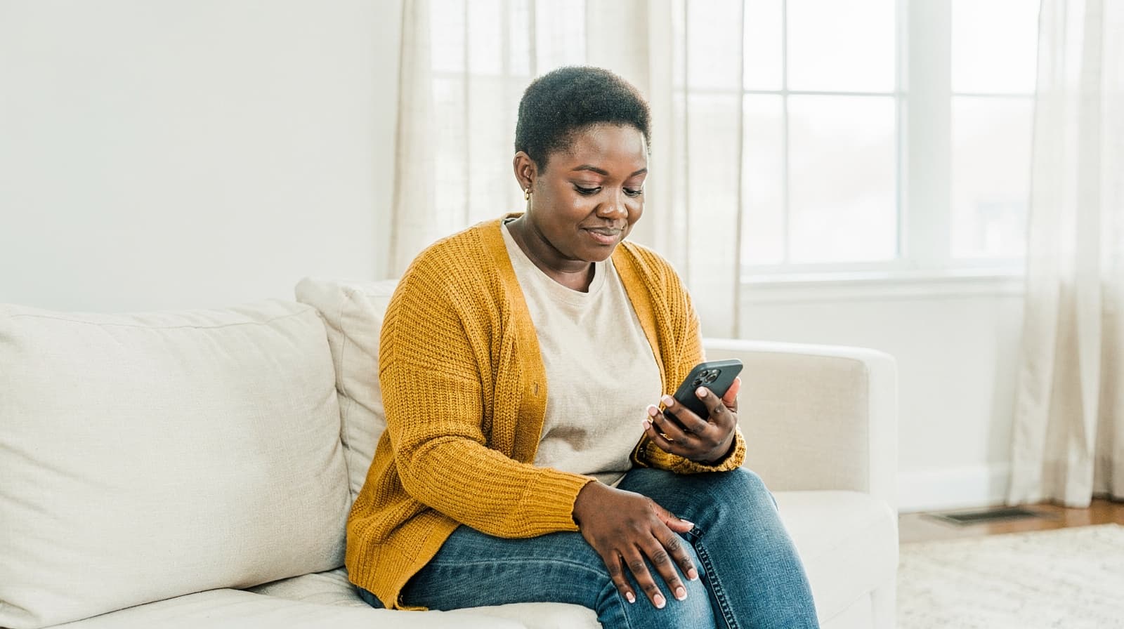 A woman in a yellow cardigan sits on a couch using a smartphone in a bright living room.
