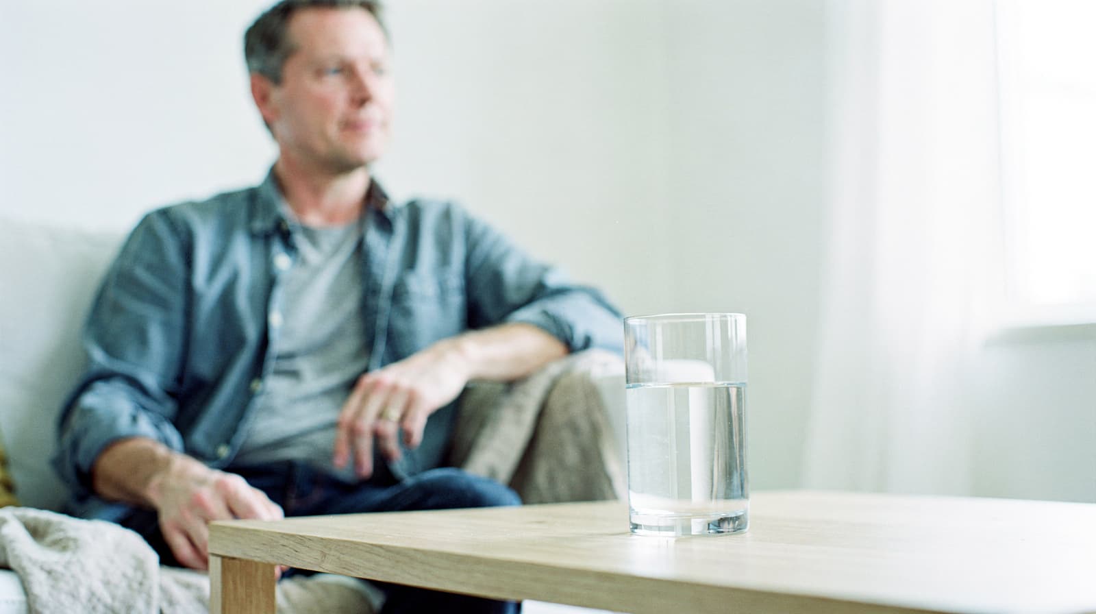 A man sits on a couch in a bright room with a glass of water on a wooden table in the foreground.