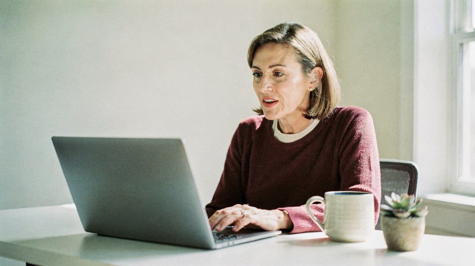 A woman sits at a desk using a laptop with a mug and a small plant beside her.