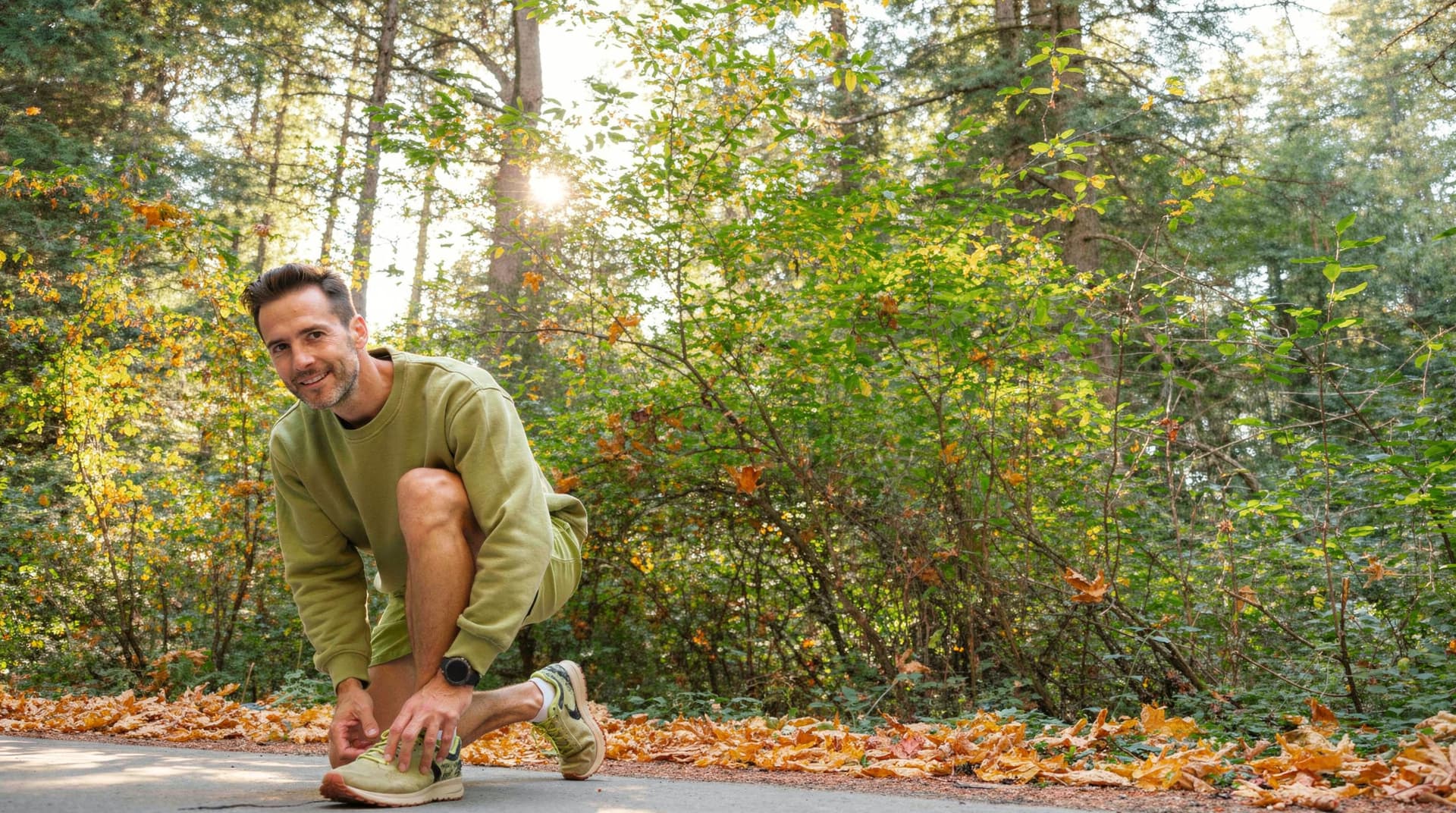A man kneels to tie his shoe on a forest path surrounded by autumn leaves and trees.