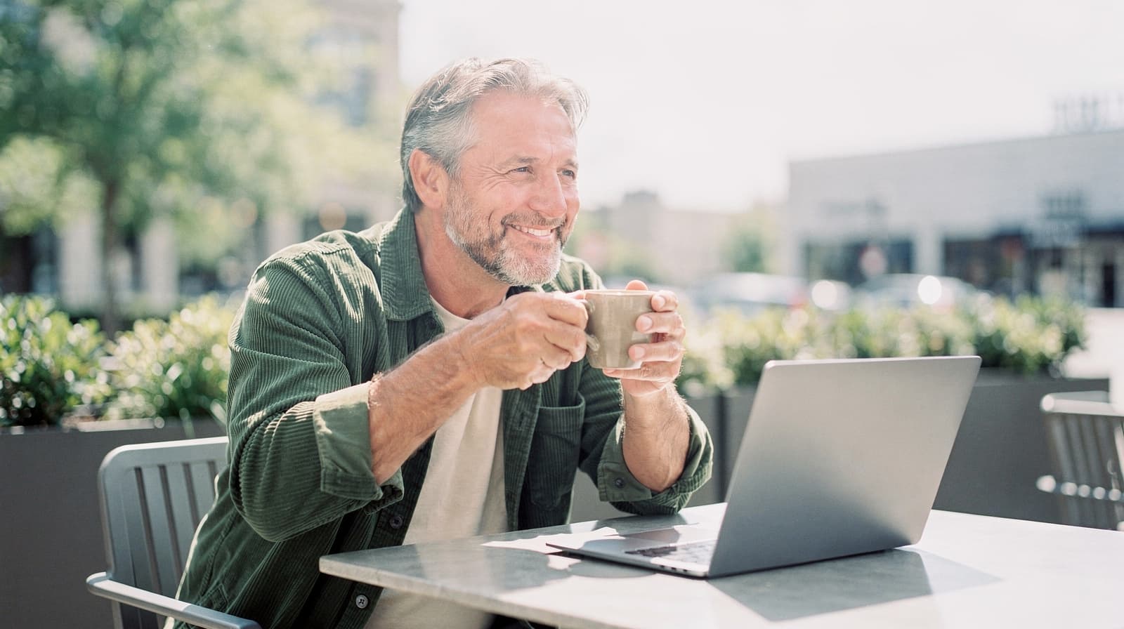 A man sitting at an outdoor cafe table smiles while holding a mug and looking at a laptop.