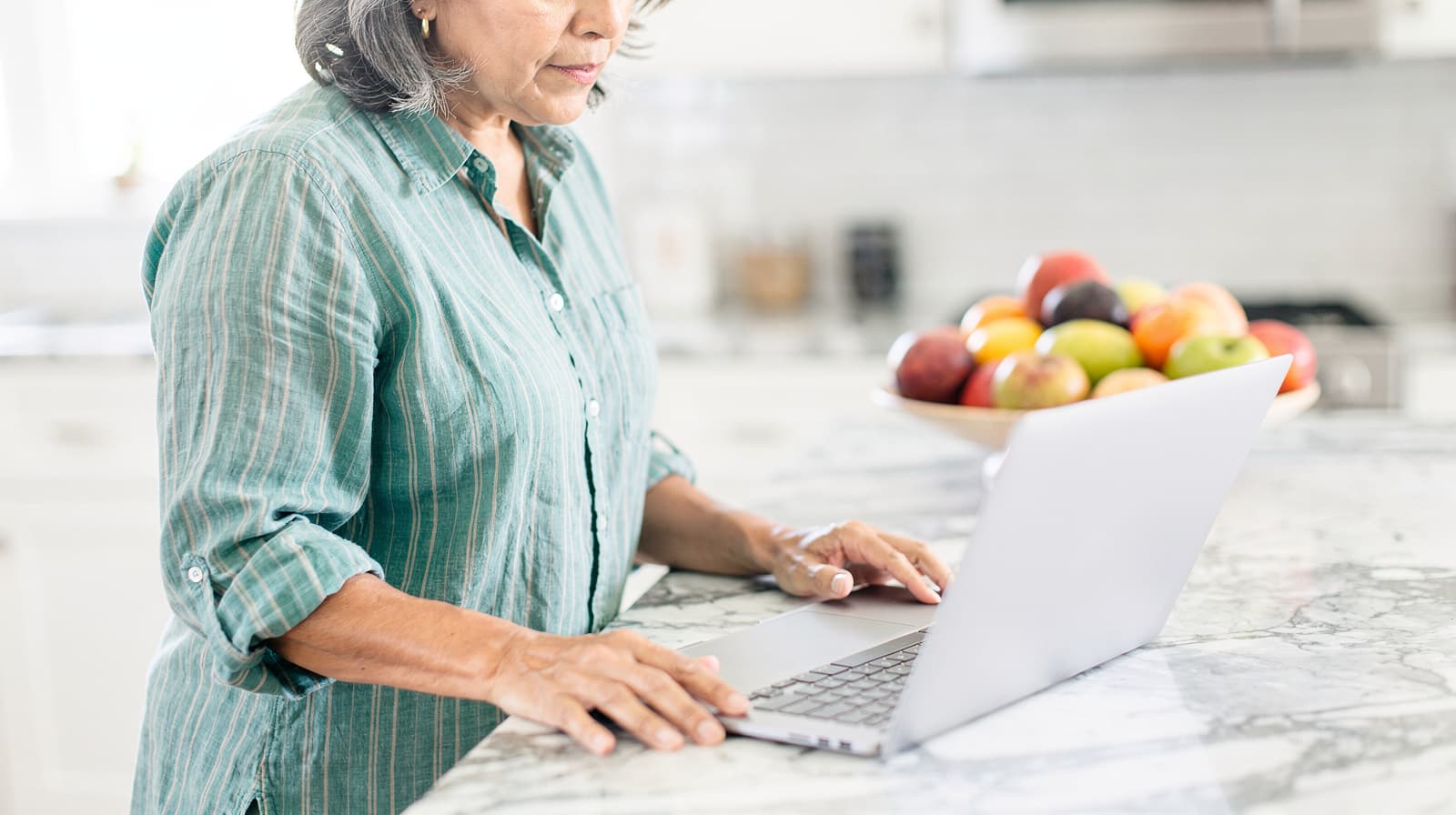 A woman in a striped shirt uses a laptop on a marble kitchen counter with a bowl of fruit nearby.