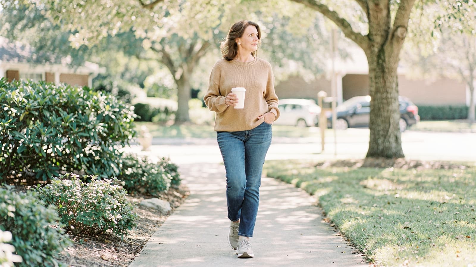 A woman walks on a sidewalk holding a coffee cup in a leafy suburban neighborhood.