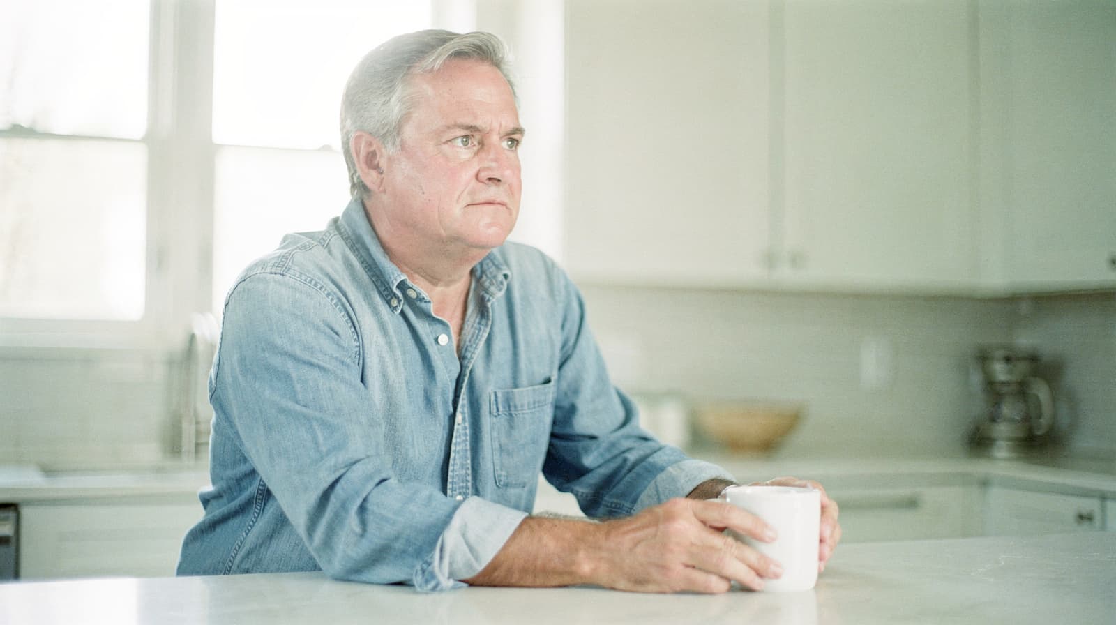 A man in a denim shirt sits at a kitchen counter holding a white mug.