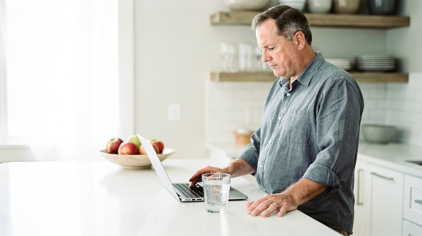 A man stands at a kitchen counter using a laptop with a glass of water nearby.