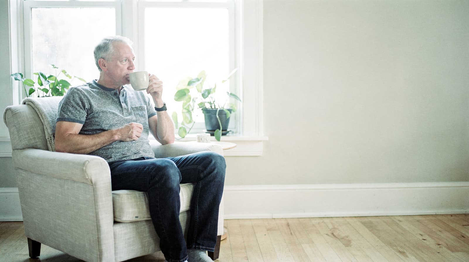 A man sits in an armchair by a window, drinking from a mug, with potted plants on the windowsill.