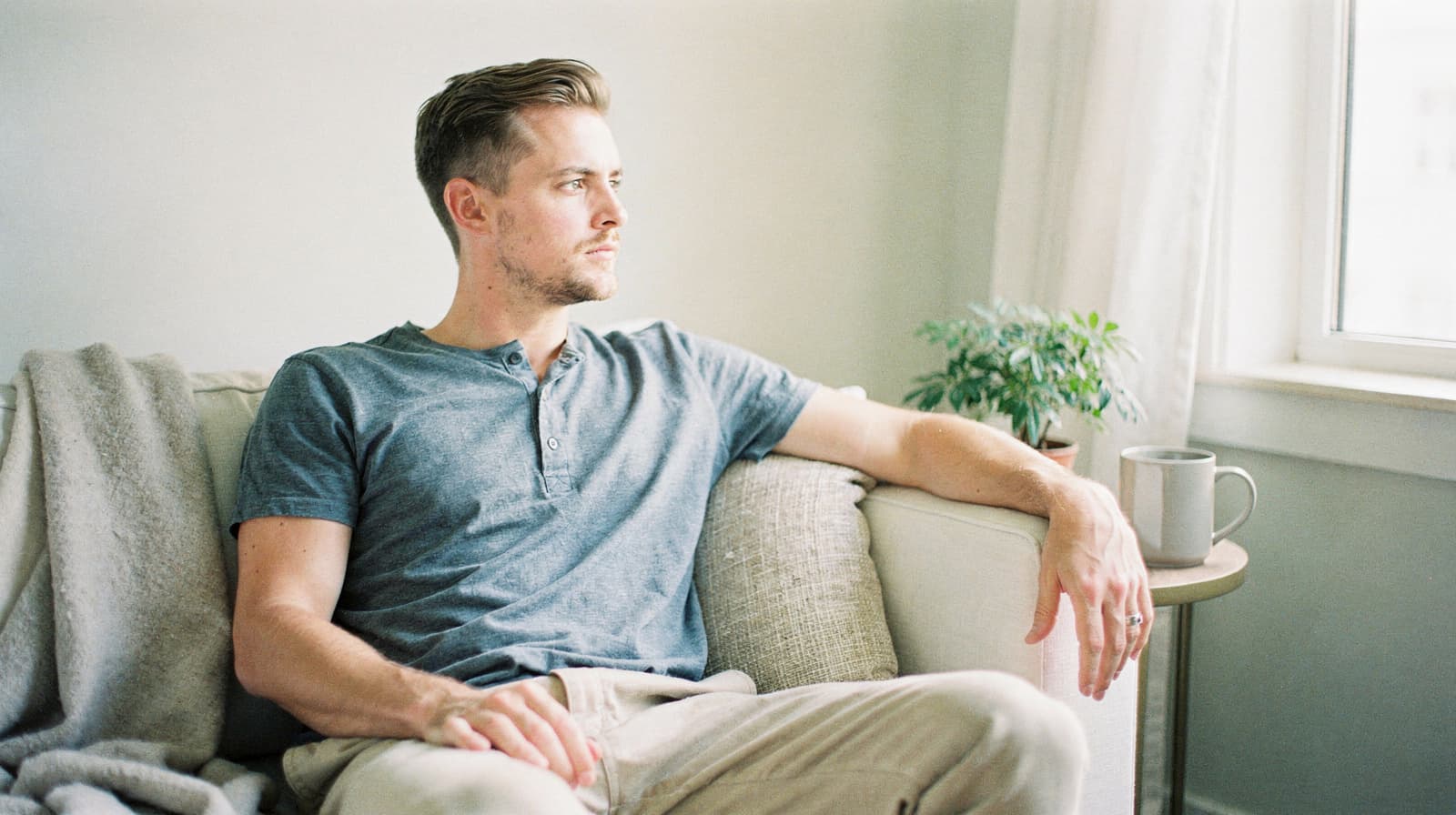 A man sits on a couch looking out a window with a mug and a small plant nearby.