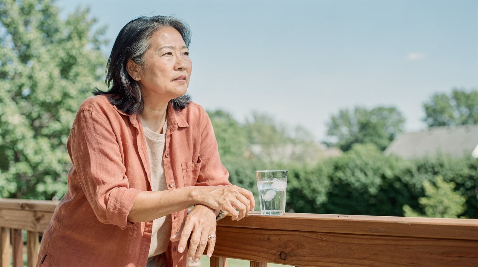 A woman in a red shirt stands on a wooden deck, holding a glass of water, with trees and a clear sky in the background.