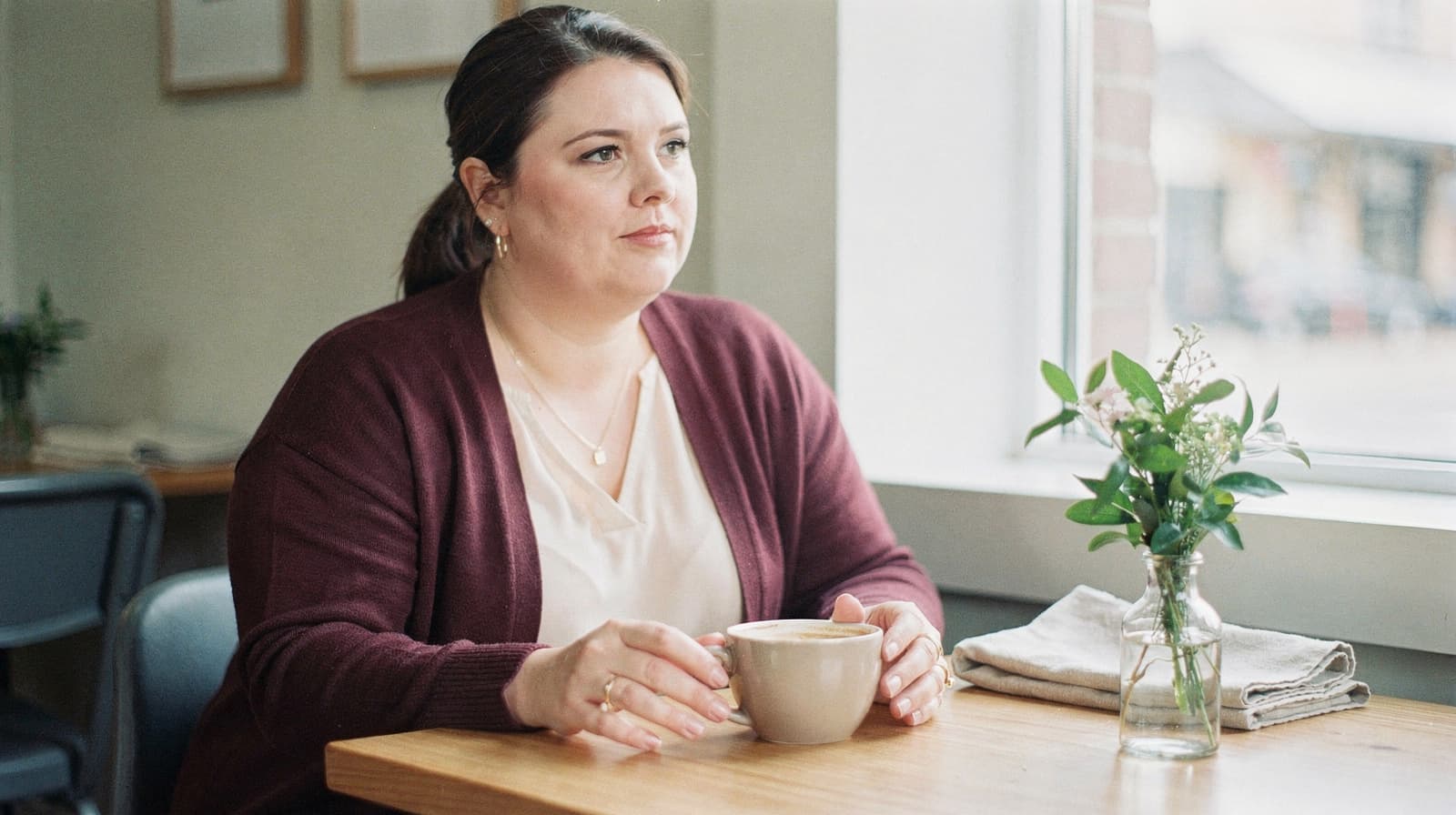 A woman sits at a wooden table in a cafe, holding a cup and looking out the window.