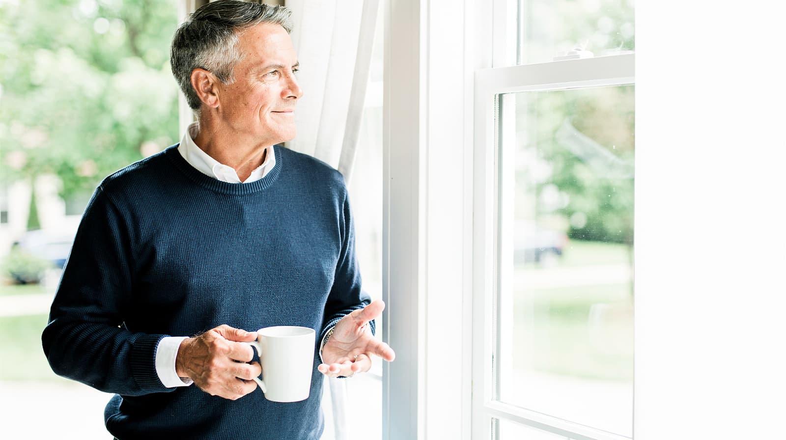 A man holding a mug looks out a window in a bright room.