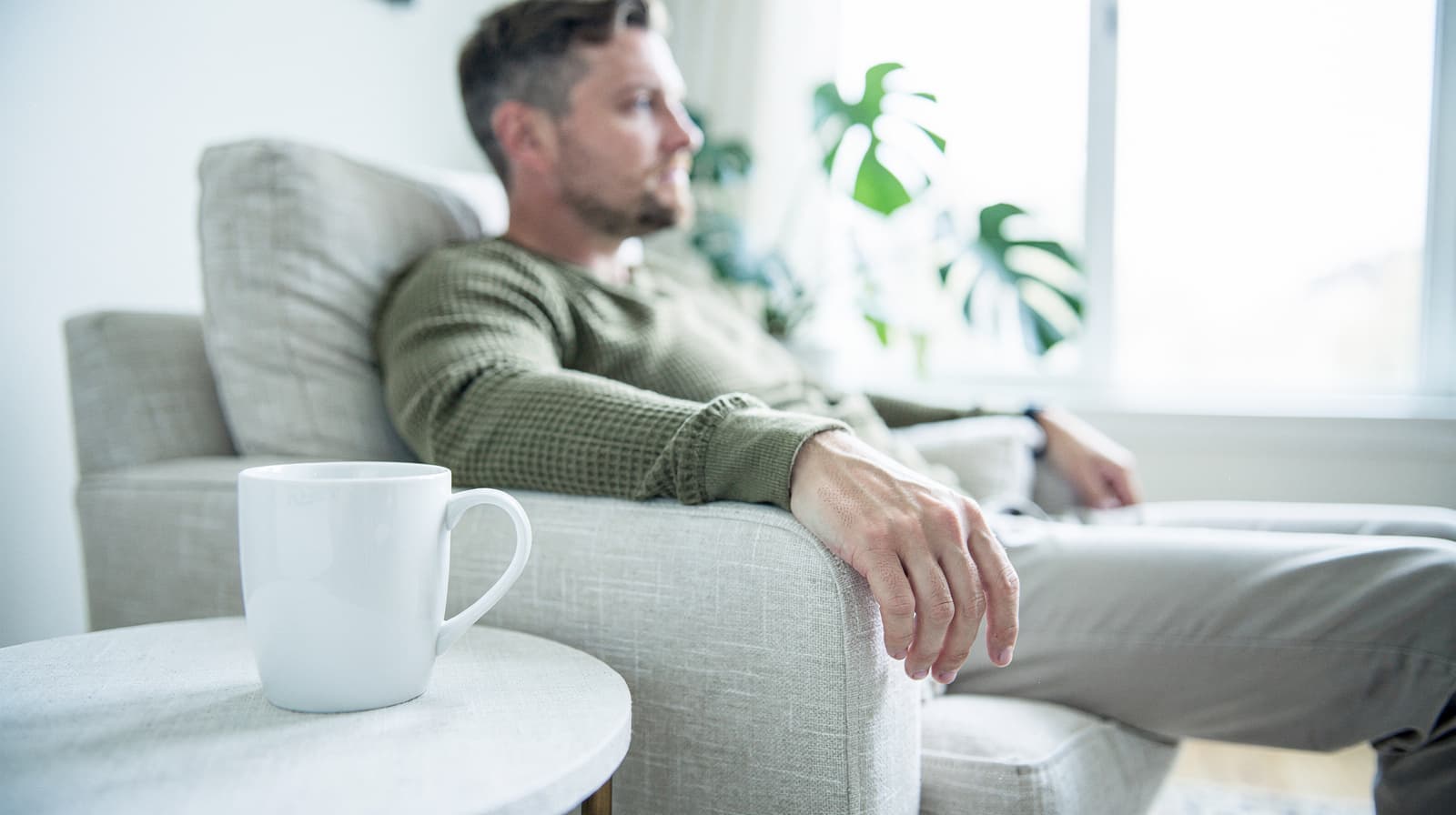 A man sits on a light-colored armchair in a bright room with a white mug on a nearby table.