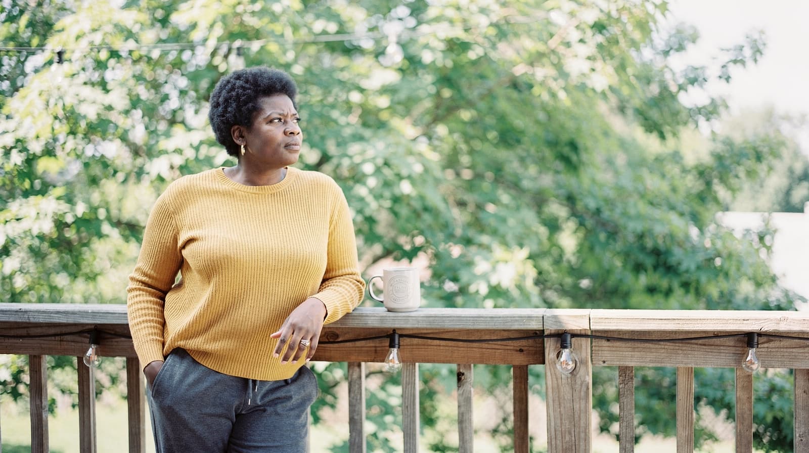 A person in a yellow sweater stands on a wooden deck with trees in the background, holding a mug.