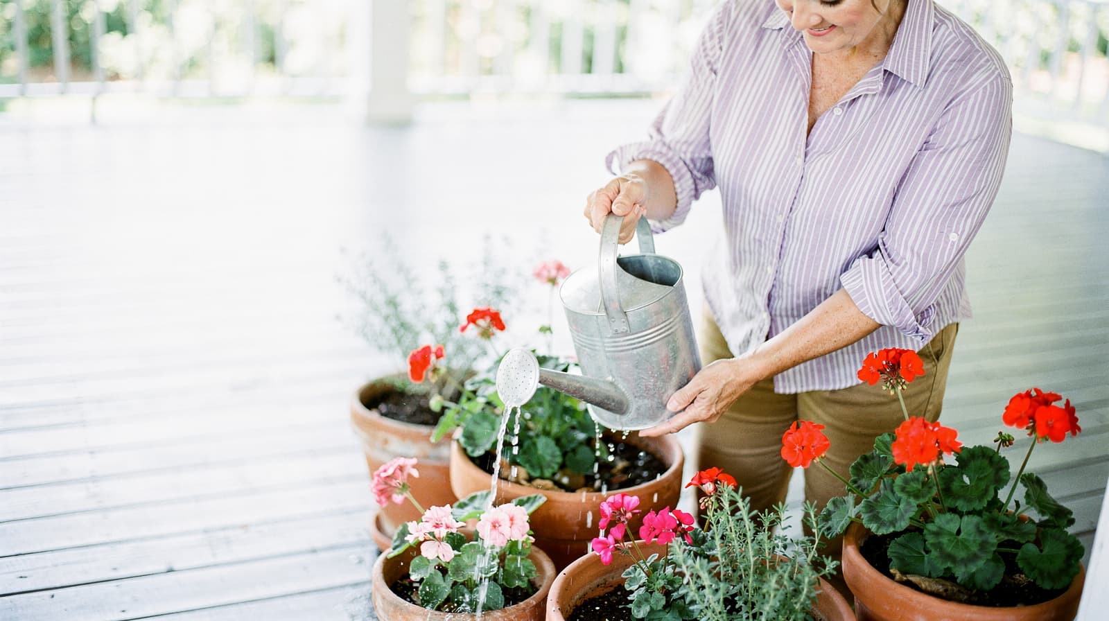 A woman waters potted flowers on a wooden porch.