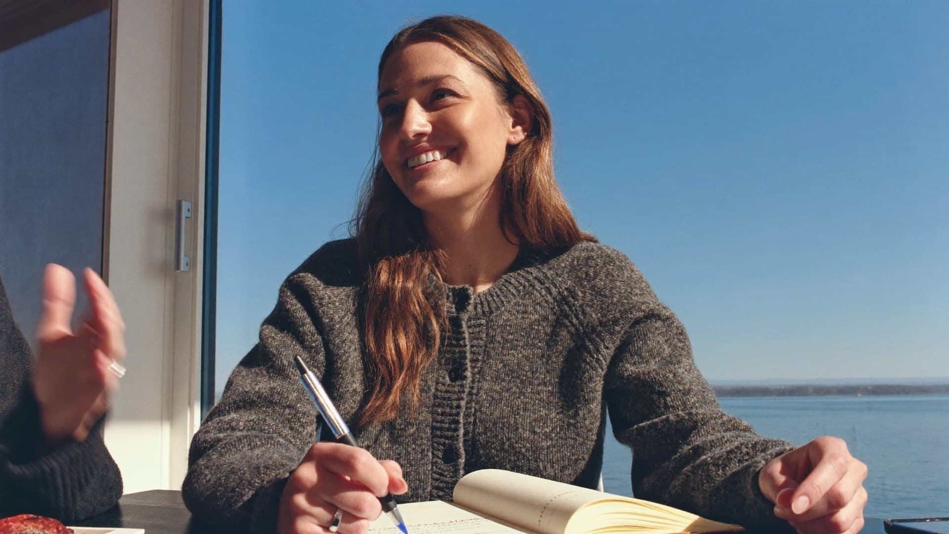 A woman writing at a table by a window during a conversation.