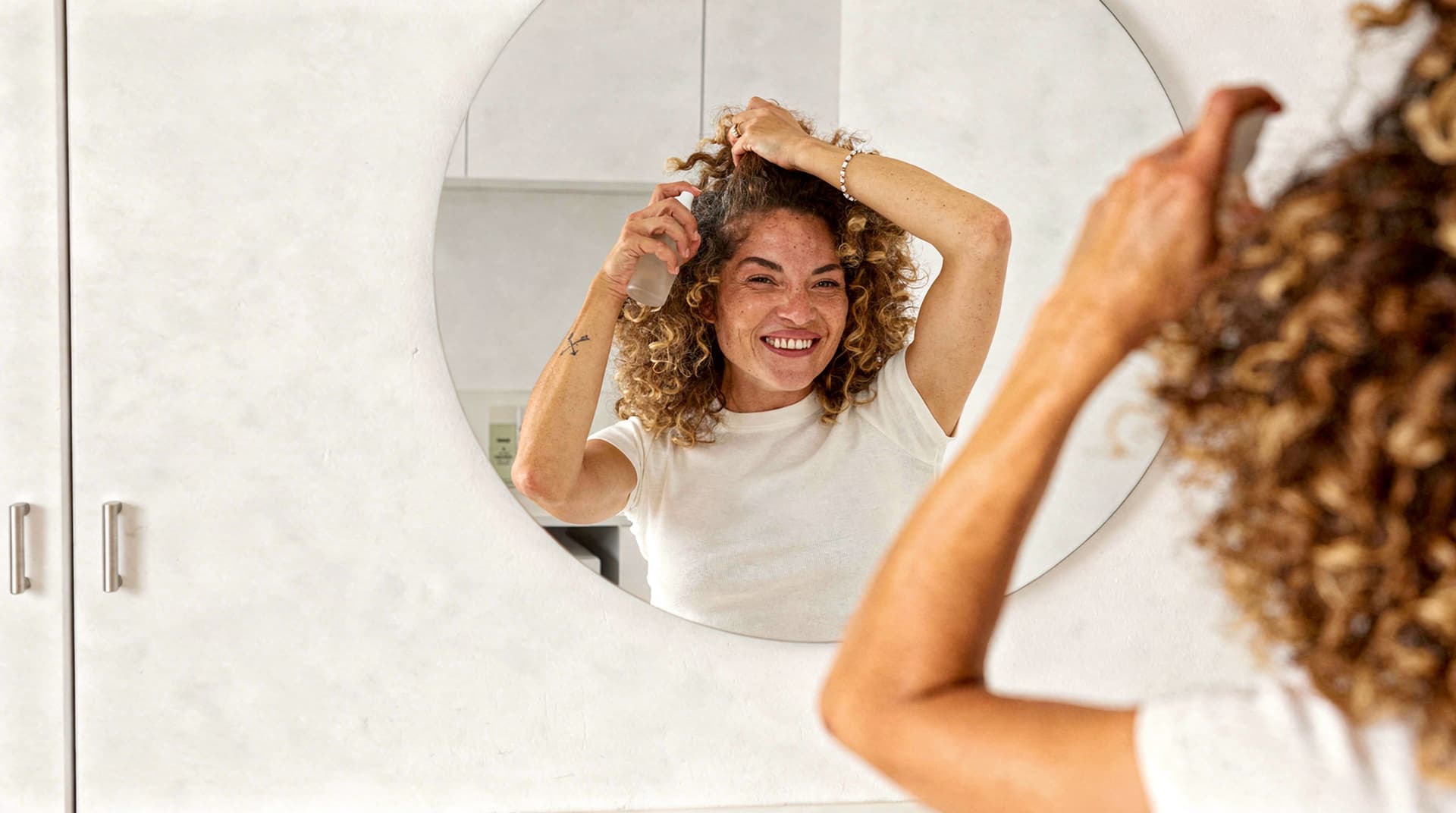 A woman with curly hair smiles while styling her hair in front of a round mirror in a bathroom.