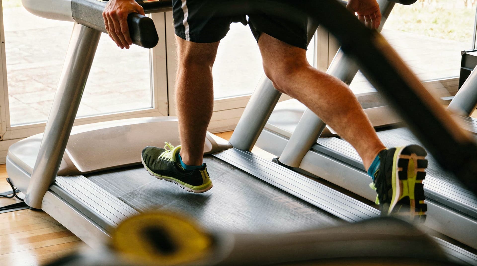 A person wearing athletic shoes runs on a treadmill indoors near a large window.