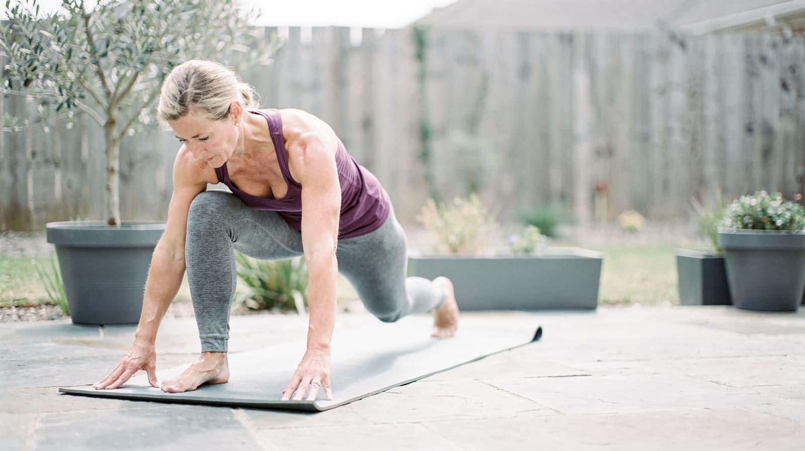 A woman in athletic wear performs a lunge on a yoga mat in a backyard patio.