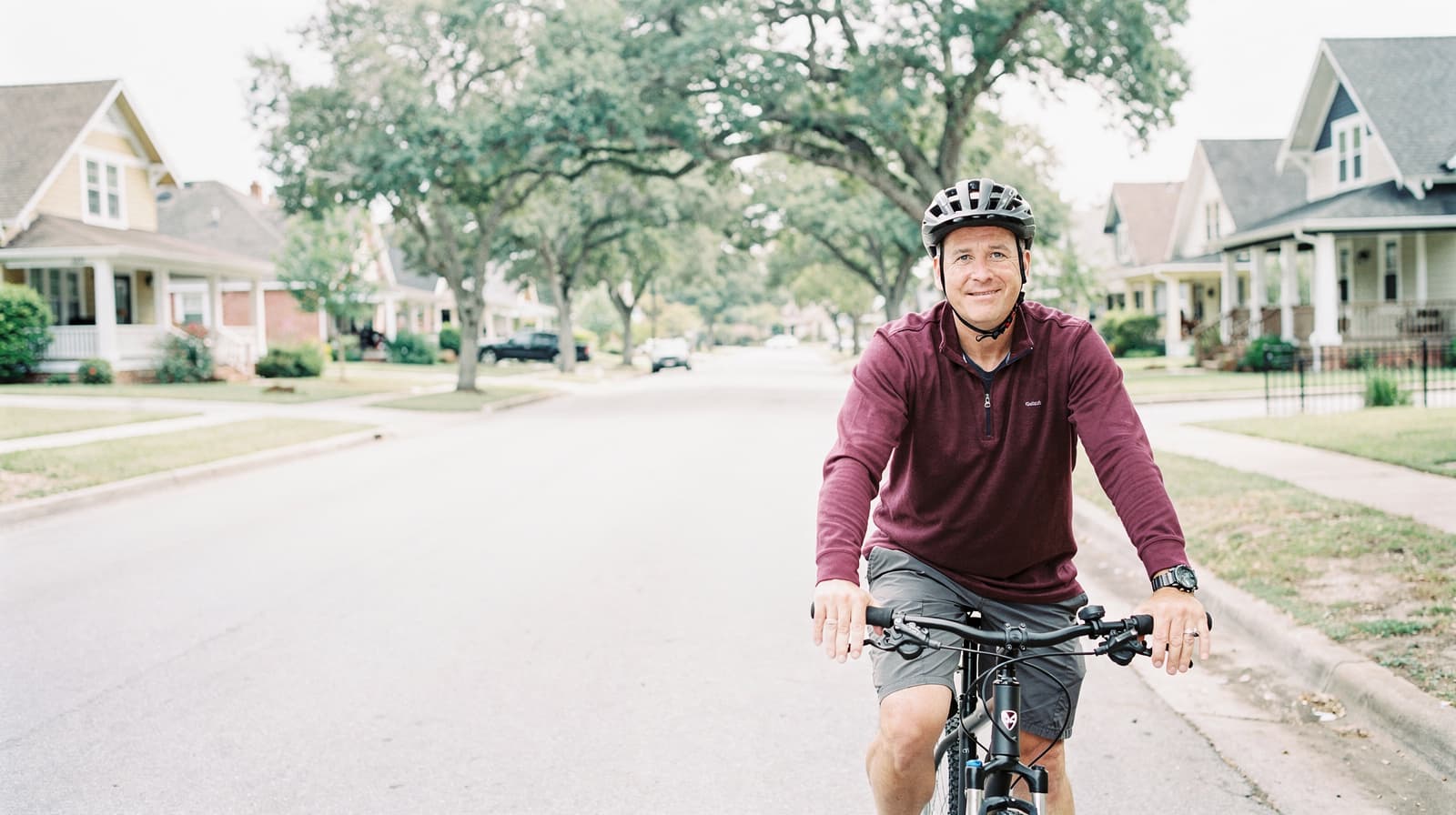 A man wearing a helmet rides a bicycle down a suburban street lined with houses and trees.