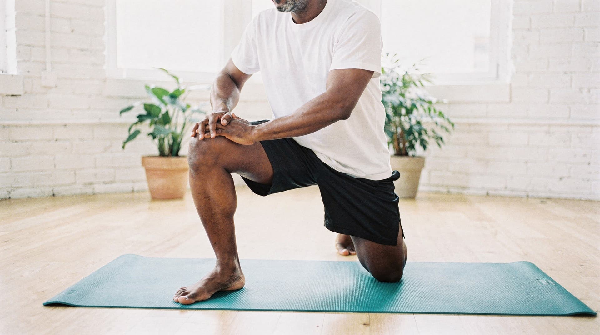 A man in a white shirt and black shorts kneels on a yoga mat in a bright room with plants.